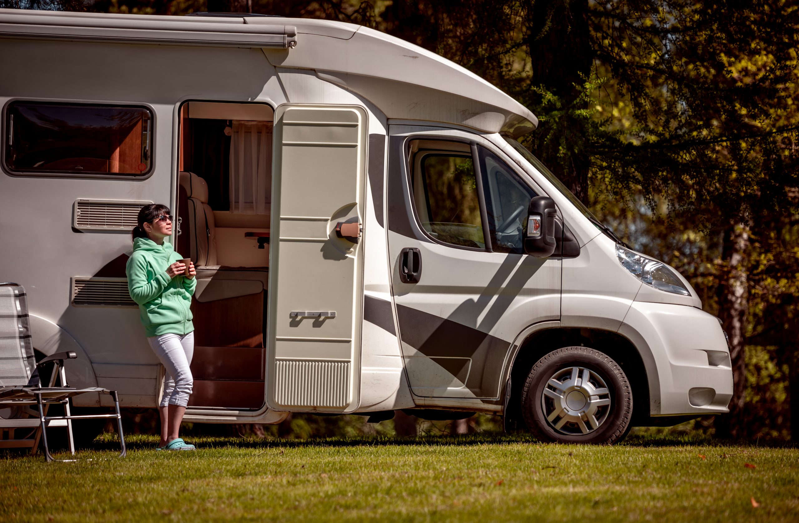 Woman is standing with a mug of coffee near the camper RV. Eine Frau mit einem Becher Kaffee steht in die Tür ihres Wohnmobils gelehnt