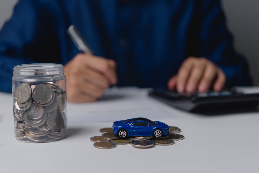 Auf einem Tisch steht ein Einmachglas gefüllt mit Geldmünzen, daneben ein kleines blaues Spielzeugauto auf ein paar weiteren Münzen