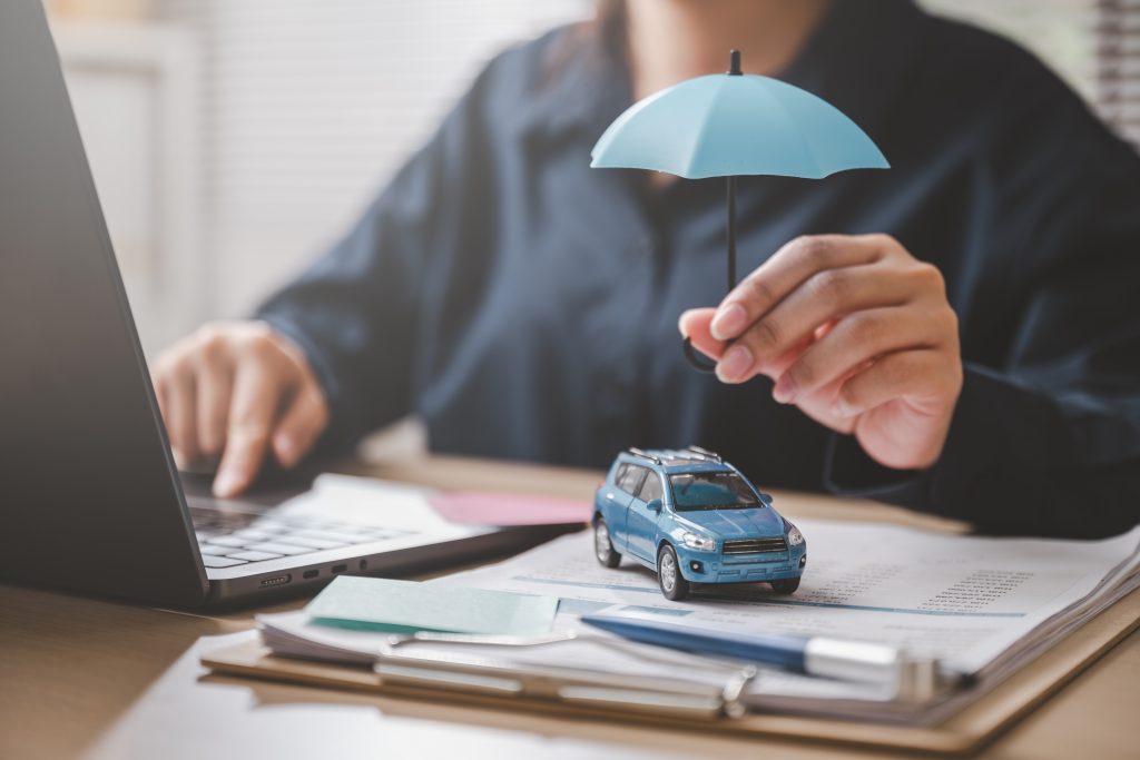 Auf einem Schreibtisch liegen Versicherungsunterlagen. Darauf ein kleines blaues Spielzeugauto, über das ein Puppen-Schirm gehalten wird.